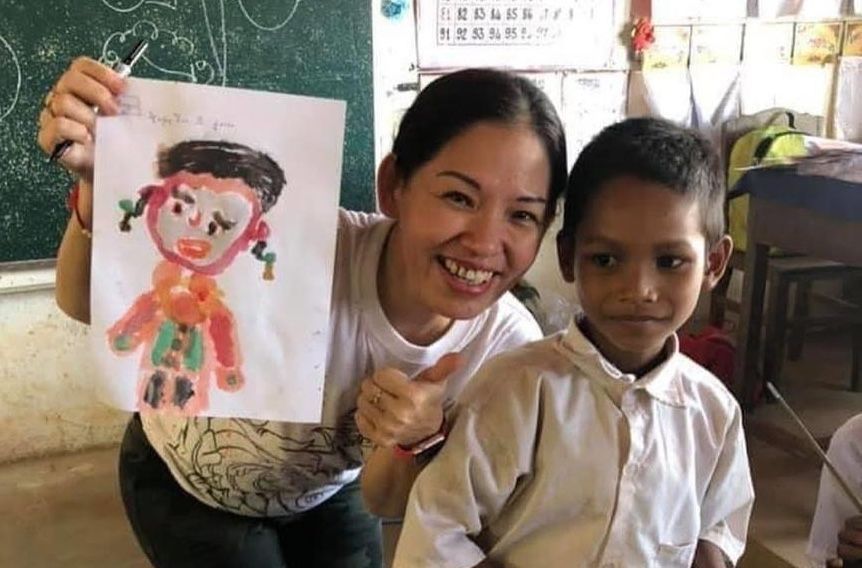 Honey Khor smiling beside a schoolboy in Cambodia, holding up his painting
